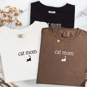 May include: Three t-shirts in black, white, and brown, each with the words "cat mom" printed on the front. A gold necklace rests on the brown shirt. The shirts are made of a soft material.