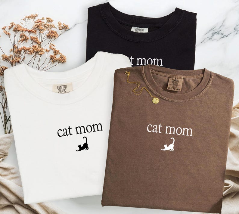 May include: Three t-shirts in black, white, and brown, each with the words "cat mom" printed on the front. A gold necklace rests on the brown shirt. The shirts are made of a soft material.