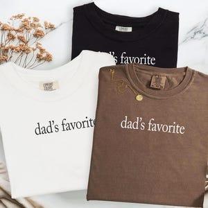 May include: Three t-shirts in black, white, and brown, each with the text "dad's favorite" in white. The white t-shirt has a Comfort Colors tag. A gold necklace is draped on the brown shirt. The shirts are on a marble surface.