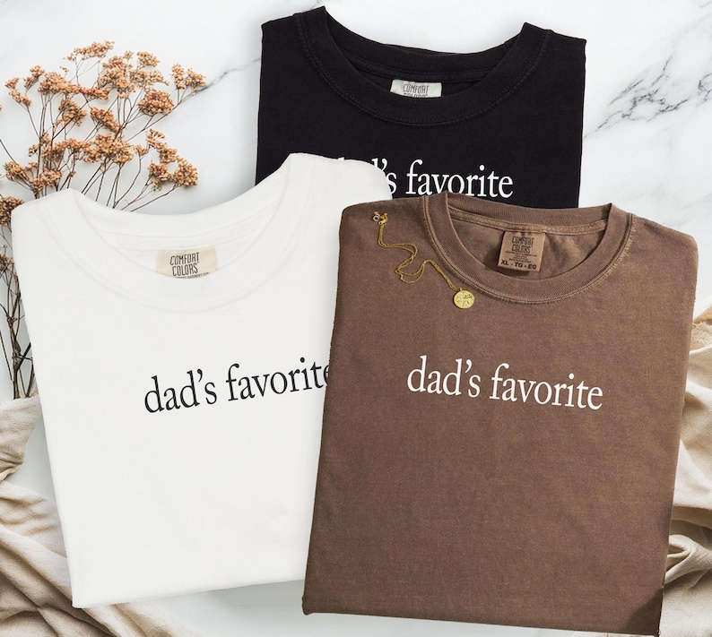 May include: Three t-shirts in black, white, and brown, each with the text "dad's favorite" in white. The white t-shirt has a Comfort Colors tag. A gold necklace is draped on the brown shirt. The shirts are on a marble surface.
