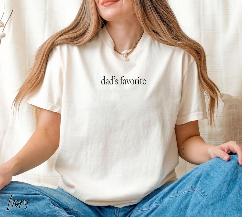 May include: Ivory-colored t-shirt with the text "dad's favorite" in black. The shirt has short sleeves and a crew neck. The model is wearing blue jeans.