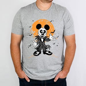 May include: A grey t-shirt featuring a black and white illustration of Mickey Mouse dressed as Jack Skellington from The Nightmare Before Christmas. The illustration depicts Mickey Mouse wearing a black and white striped suit, a black bow tie, and a stitched-up mouth. The background is a dark orange moon with black bats flying around it.