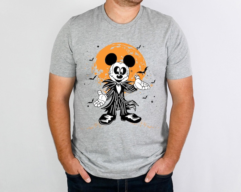 May include: A grey t-shirt featuring a black and white illustration of Mickey Mouse dressed as Jack Skellington from The Nightmare Before Christmas. The illustration depicts Mickey Mouse wearing a black and white striped suit, a black bow tie, and a stitched-up mouth. The background is a dark orange moon with black bats flying around it.