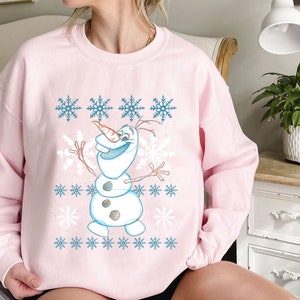 May include: Pink sweatshirt with a cartoon snowman design and a snowflake pattern. The snowman is wearing a blue scarf and has a carrot nose.