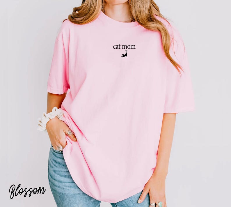 May include: A light pink t-shirt with the text "cat mom" and a small black cat graphic. The shirt is a loose fit and is paired with blue jeans. The word "Blossom" is written in cursive in the bottom left corner.