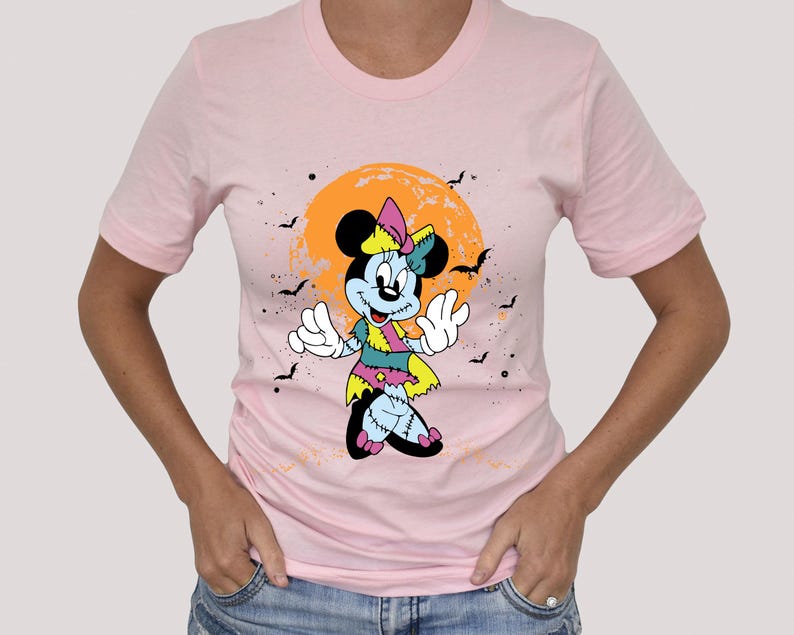 May include: Pink t-shirt featuring a cartoon image of a zombie Minnie Mouse in front of a yellow and orange moon. The image has a Halloween theme.