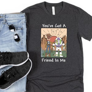 May include: Dark gray t-shirt with a graphic featuring Woody, Buzz Lightyear, and the text "You've Got A Friend In Me." The shirt is paired with blue jeans, black sneakers, and sunglasses.