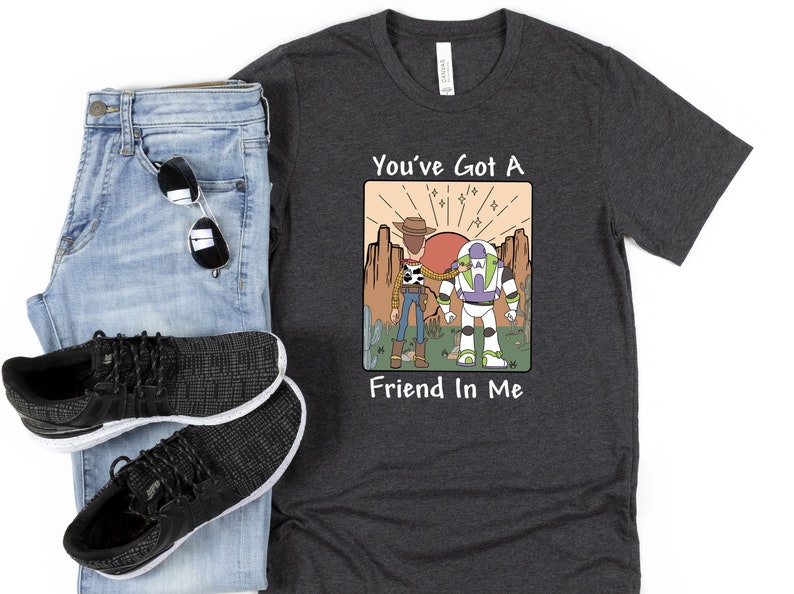 May include: Dark gray t-shirt with a graphic featuring Woody, Buzz Lightyear, and the text "You've Got A Friend In Me." The shirt is paired with blue jeans, black sneakers, and sunglasses.