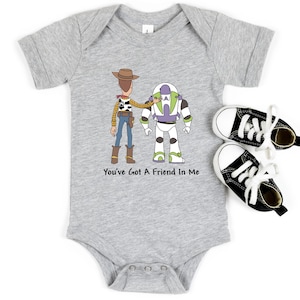 Puede incluir: Un body de beb&eacute; gris con una imagen de dibujos animados de Woody y Buzz Lightyear de Toy Story. El body tiene el texto "You've Got A Friend In Me" impreso en &eacute;l.