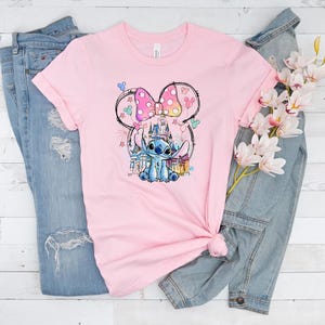 K&ouml;nnte beinhalten: Ein hellrosa T-Shirt mit einer farbenfrohen Grafik von Stitch aus Lilo & Stitch, mit einer Minnie Mouse-inspirierten Schleife und einem Schloss im Hintergrund. Das Shirt liegt auf einer blauen Jeans und einer Jeansjacke.
