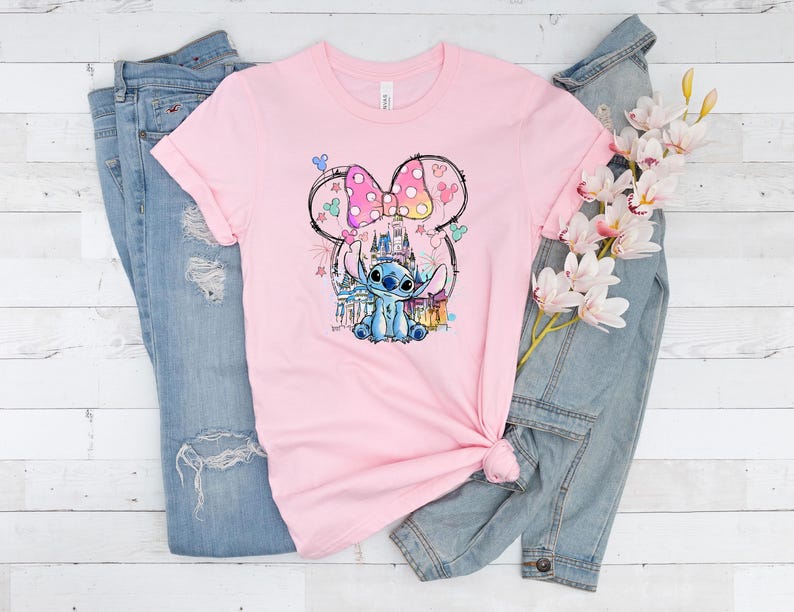 K&ouml;nnte beinhalten: Ein hellrosa T-Shirt mit einer farbenfrohen Grafik von Stitch aus Lilo & Stitch, mit einer Minnie Mouse-inspirierten Schleife und einem Schloss im Hintergrund. Das Shirt liegt auf einer blauen Jeans und einer Jeansjacke.