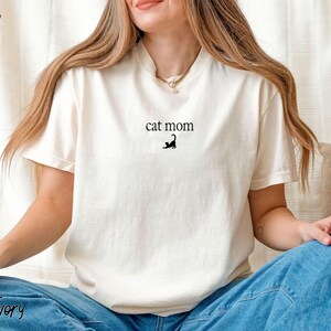 May include: Ivory-colored t-shirt with the text "cat mom" and a black cat silhouette. The shirt is made of a soft material and has a classic crew neck design. The shirt is a casual wear item.