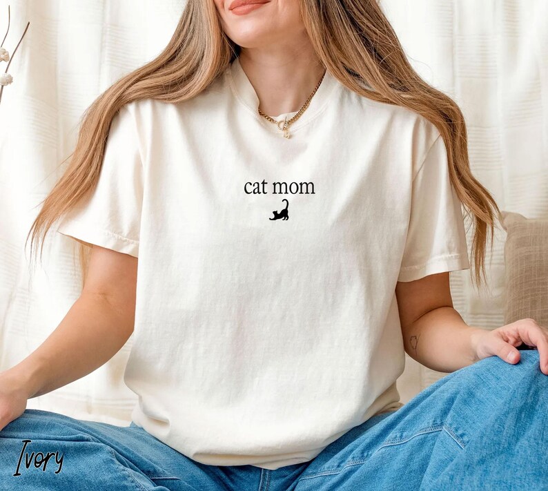 May include: Ivory-colored t-shirt with the text "cat mom" and a black cat silhouette. The shirt is made of a soft material and has a classic crew neck design. The shirt is a casual wear item.