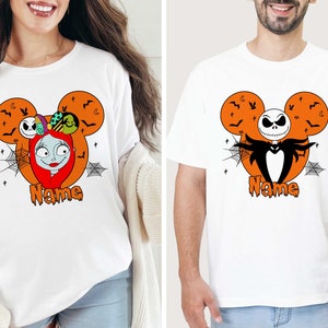 Op de afbeelding: Twee witte T-shirts met een Halloween Mickey Mouse-ontwerp. E&eacute;n shirt heeft Sally van The Nightmare Before Christmas en de andere heeft Jack Skellington. Beide shirts hebben de tekst "Name" onder het ontwerp gedrukt.