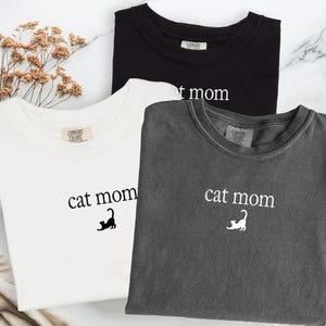 May include: Three Comfort Colors t-shirts in black, white, and charcoal gray. Each shirt features the text "cat mom" with a cat graphic. The shirts are laid flat on a marble-like surface.
