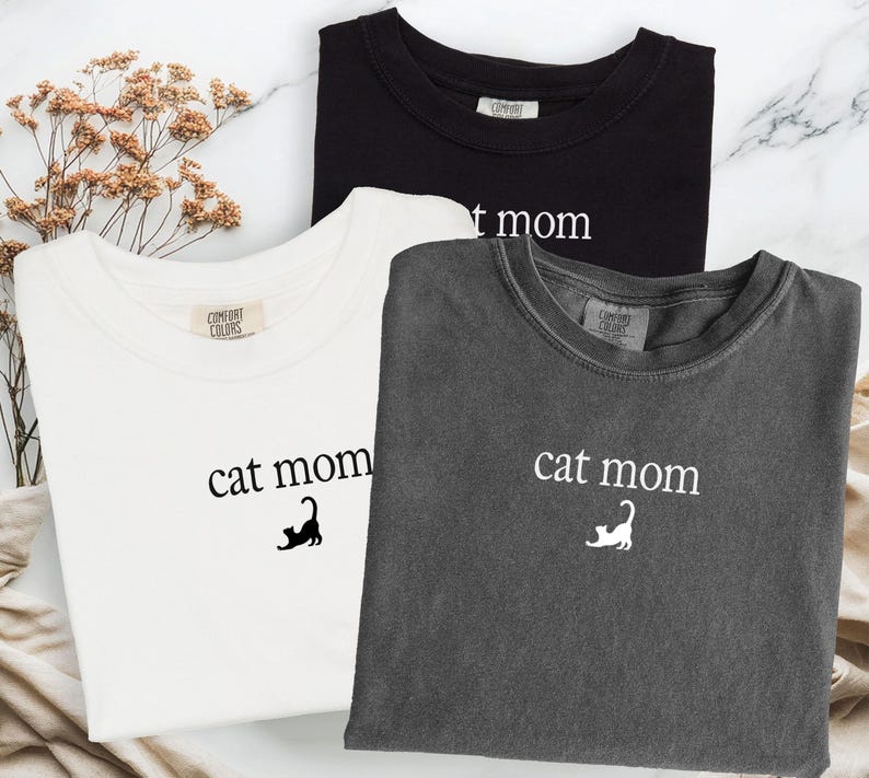 May include: Three Comfort Colors t-shirts in black, white, and charcoal gray. Each shirt features the text "cat mom" with a cat graphic. The shirts are laid flat on a marble-like surface.