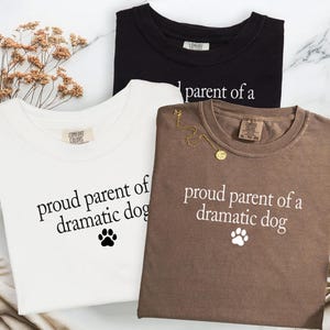 May include: Three t-shirts in black, white, and brown, each with the phrase "proud parent of a dramatic dog" printed in white. A paw print graphic is below the text. The white shirt has a Comfort Colors tag.