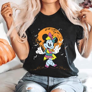 May include: Black t-shirt featuring a graphic of a stitched-up Mickey Mouse character in front of a jack-o-lantern. The character is wearing a pink and green dress and has a stitched-up nose.