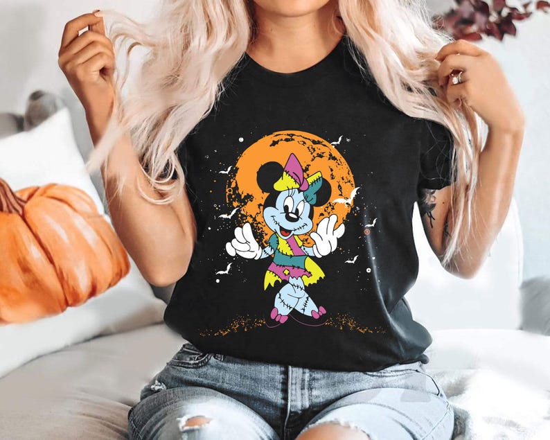 May include: Black t-shirt featuring a graphic of a stitched-up Mickey Mouse character in front of a jack-o-lantern. The character is wearing a pink and green dress and has a stitched-up nose.