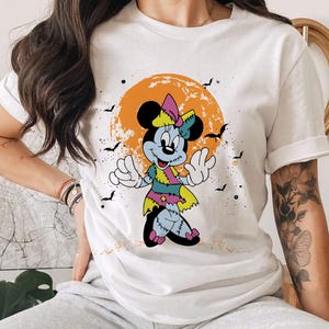 May include: White t-shirt featuring a graphic of a zombie Minnie Mouse in a Halloween setting. The graphic depicts a cartoon Minnie Mouse with green and purple clothing, stitched seams, and a bright orange background.