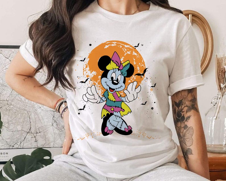 May include: White t-shirt featuring a graphic of a zombie Minnie Mouse in a Halloween setting. The graphic depicts a cartoon Minnie Mouse with green and purple clothing, stitched seams, and a bright orange background.