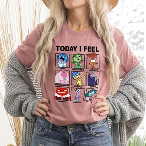 Pu&ograve; includere: Una maglietta rosa con una grafica dei personaggi del film Inside Out. La grafica dice "Oggi mi sento" e ha una griglia di nove quadrati con diverse emozioni. Ogni quadrato ha un personaggio del film e il nome dell'emozione. Le emozioni sono: Triste, Disgustato, Ansioso, Imbarcato, Gioioso, Annoiato, Arrabbiato, Paura e Deluso.