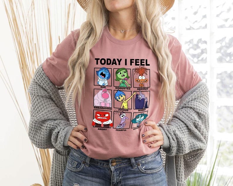 Pu&ograve; includere: Una maglietta rosa con una grafica dei personaggi del film Inside Out. La grafica dice "Oggi mi sento" e ha una griglia di nove quadrati con diverse emozioni. Ogni quadrato ha un personaggio del film e il nome dell'emozione. Le emozioni sono: Triste, Disgustato, Ansioso, Imbarcato, Gioioso, Annoiato, Arrabbiato, Paura e Deluso.