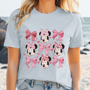 以下が含まれることがあります： 水色のTシャツで、水玉模様のリボンをつけたミニーマウスの顔、無地のピンクのリボン、小さなピンクのハートと星がグリッド状に配置されたグラフィックパターンが前面にプリントされています。