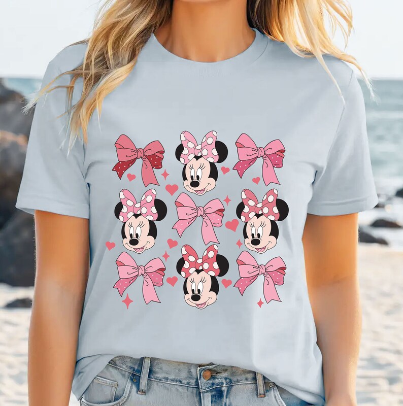 以下が含まれることがあります： 水色のTシャツで、水玉模様のリボンをつけたミニーマウスの顔、無地のピンクのリボン、小さなピンクのハートと星がグリッド状に配置されたグラフィックパターンが前面にプリントされています。