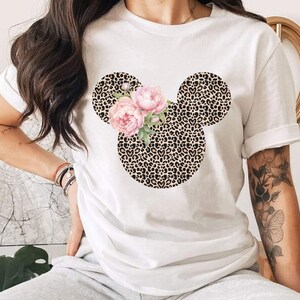 May include: White t-shirt featuring a Mickey Mouse head design with a leopard print pattern. The ears are leopard print, and the face is adorned with pink flowers. A stylish and unique graphic tee.