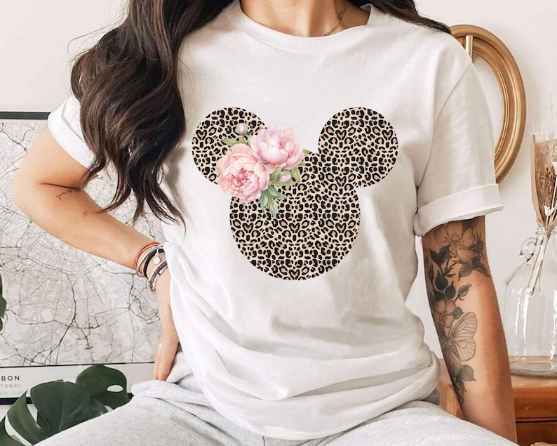 May include: White t-shirt featuring a Mickey Mouse head design with a leopard print pattern. The ears are leopard print, and the face is adorned with pink flowers. A stylish and unique graphic tee.