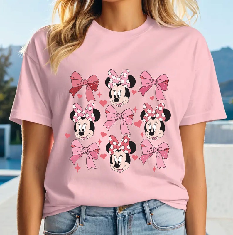 以下が含まれることがあります： ライトピンクの半袖Tシャツ。前面には、水玉模様のリボンや無地のピンクのリボンをつけたミニーマウスの顔、小さな赤いハートや星の形がグリッド状に配置されたグラフィックパターンがプリントされています。