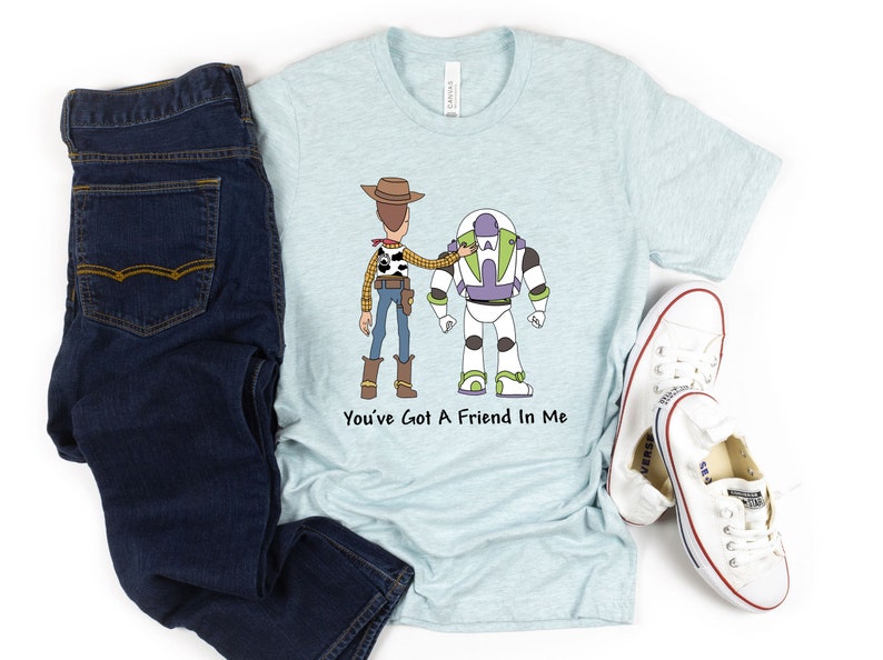 Puede incluir: Una camiseta azul claro con una imagen de dibujos animados de Woody y Buzz Lightyear de Toy Story. La camiseta tiene el texto "You've Got A Friend In Me" impreso en ella.