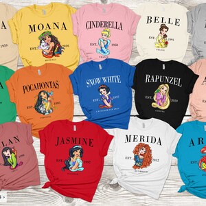 May include: A collection of 12 t-shirts featuring different Disney Princesses. Each shirt has a different princess, her name, and the year she was established. The shirts are in various colors, including pink, blue, green, red, orange, and white.