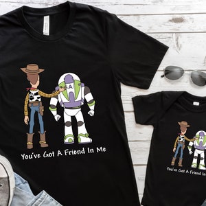 Puede incluir: Una camiseta negra con una imagen de dibujos animados de Woody y Buzz Lightyear de Toy Story. La camiseta tiene el texto "You've Got A Friend In Me" impreso en ella. Tambi&eacute;n se muestra un body negro a juego para beb&eacute; con la misma imagen y texto.