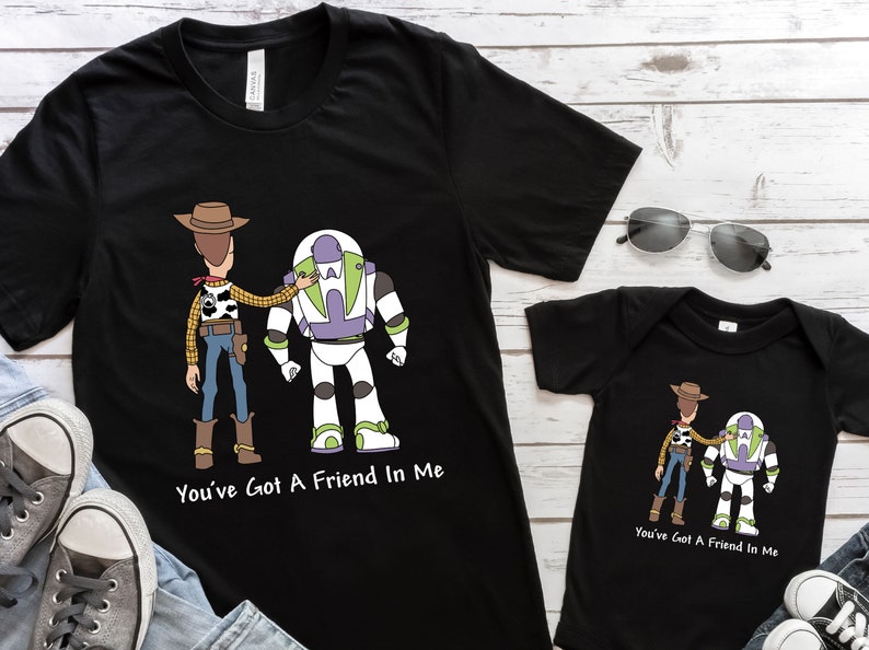 Puede incluir: Una camiseta negra con una imagen de dibujos animados de Woody y Buzz Lightyear de Toy Story. La camiseta tiene el texto "You've Got A Friend In Me" impreso en ella. Tambi&eacute;n se muestra un body negro a juego para beb&eacute; con la misma imagen y texto.