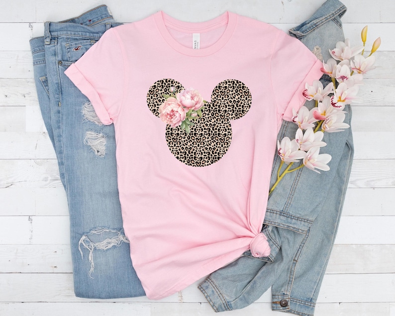 May include: A light pink t-shirt featuring a Mickey Mouse head design with a leopard print pattern and pink floral accents. The shirt is styled with light blue denim jeans and a sprig of white and pink flowers.