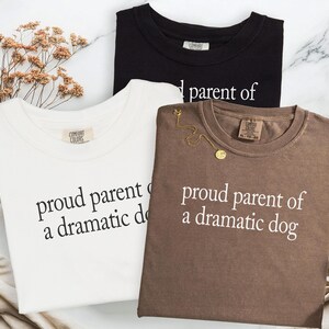 May include: Three t-shirts in black, white, and brown, each with the phrase "proud parent of a dramatic dog" printed in white. The shirts are displayed on a marble surface, with a sprig of dried flowers.