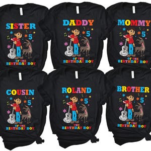 May include: Six black t-shirts with colorful text and graphics. The shirts feature a cartoon character playing a guitar with a dog beside him. The text on the shirts reads "Sister", "Daddy", "Mommy", "Cousin", "Roland", and "Brother" with the number "5" and the phrase "Of the Birthday Boy".