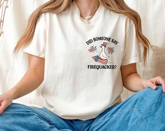 May include: A cream-colored short-sleeve t-shirt featuring a graphic on the chest of a white duck wearing a red, white, and blue top hat, riding a firecracker, surrounded by fireworks, with the text DID SOMEONE SAY FIREQUACKER?