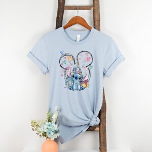 K&ouml;nnte beinhalten: Hellblaues T-Shirt mit einer Grafik von Stitch aus Lilo & Stitch, der eine Krone tr&auml;gt und vor einem Schloss sitzt. Das Design wird von einer Mickey Mouse Silhouette mit Feuerwerk und Luftballons umrahmt.