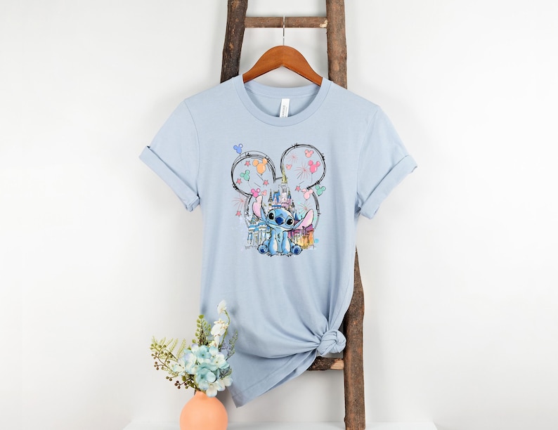 K&ouml;nnte beinhalten: Hellblaues T-Shirt mit einer Grafik von Stitch aus Lilo & Stitch, der eine Krone tr&auml;gt und vor einem Schloss sitzt. Das Design wird von einer Mickey Mouse Silhouette mit Feuerwerk und Luftballons umrahmt.