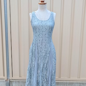 Pode incluir: Um vestido de renda azul claro em um manequim. O vestido sem mangas tem um decote redondo e um corpete ajustado que se alarga em uma saia plissada. O vestido é exibido em um suporte de metal preto com rodas, em um fundo neutro.