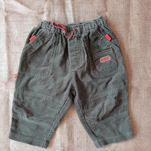 May include: Olive green corduroy shorts with a red drawstring and accents. The shorts have pockets and a small label on the right pocket. The fabric has a ribbed texture. The shorts are likely for a child.