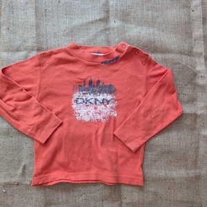 May include: Coral long-sleeved top with a crew neck and shoulder poppers. The top features a graphic print of a cityscape and the letters "DKNY" in white. The top is made of a soft material.