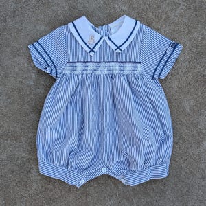 Puede incluir: Romper de bebé a rayas azules y blancas con cuello blanco y mangas cortas. El cuello tiene un ribete azul marino y un pequeño bordado de conejito. El romper tiene un corpiño smock y cierres de botón.