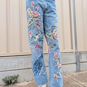 Könnte beinhalten: Hellblaue Jeans mit floralen Stickereien und Used-Details. Die Jeans zeigen bunte gestickte Blumen, Blätter und ein Pfauenmuster. Der Bund hat ein Blumenmuster. Die Jeans werden auf einer Schaufensterpuppe präsentiert.