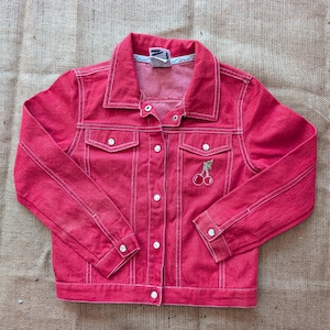 Può includere: Giacca di jeans rossa con cuciture bianche e un ricamo di ciliegie sulla tasca anteriore. La giacca ha un colletto classico, chiusure con bottoni e due tasche sul petto. La giacca è di un rosso vibrante.