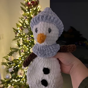 Peut inclure: Un jouet de bonhomme de neige en crochet avec un chapeau et une écharpe bleus, un corps blanc et des yeux noirs. Le bonhomme de neige est devant un sapin de Noël avec des lumières.
