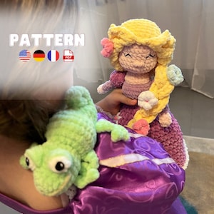 Peut inclure: Une poupée en crochet aux longs cheveux blonds portant une robe violette et tenant une fleur. La poupée est assise sur un tissu violet et blanc avec une grenouille en crochet verte.